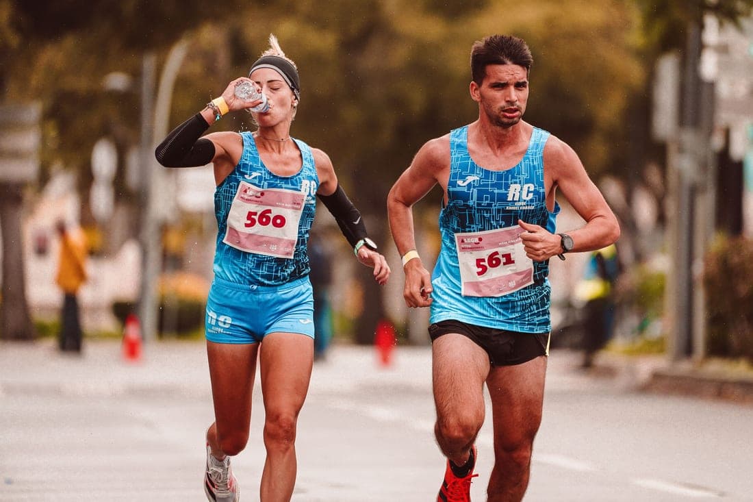 Two road-race runners, one drinking from a cup mid-race